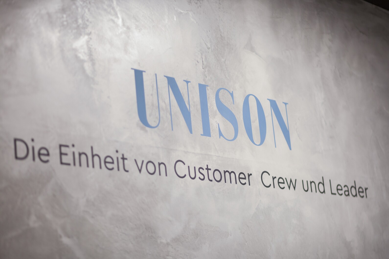 der Schriftzug "UNISON - Die Einheit von Customer Crew und Leader" auf einer Wand im Beton Look im Salon UNISON in München