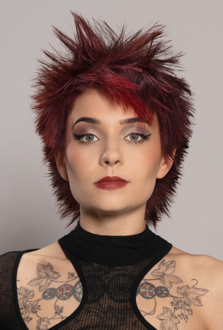 eine Frau mit einem extravaganten Pixie Kurzhaarschnitt mit asymmetrischen Stirnfransen posiert in einem schwarzen asymmetrischen Oberteil für die Kollektion Sparkle von Unison. Die Haare sind Kirschrot mit Magenta Effekten