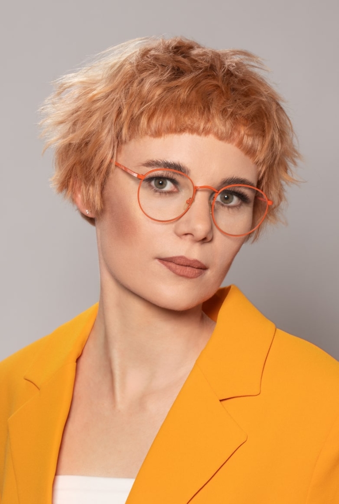 eine Frau mit einem extravaganten Pilzbob mit markantem Mikropony und Pastellkupfernen Haaren posiert in einem Orangenem Blazer für die Kollektion Sparkle von Unison