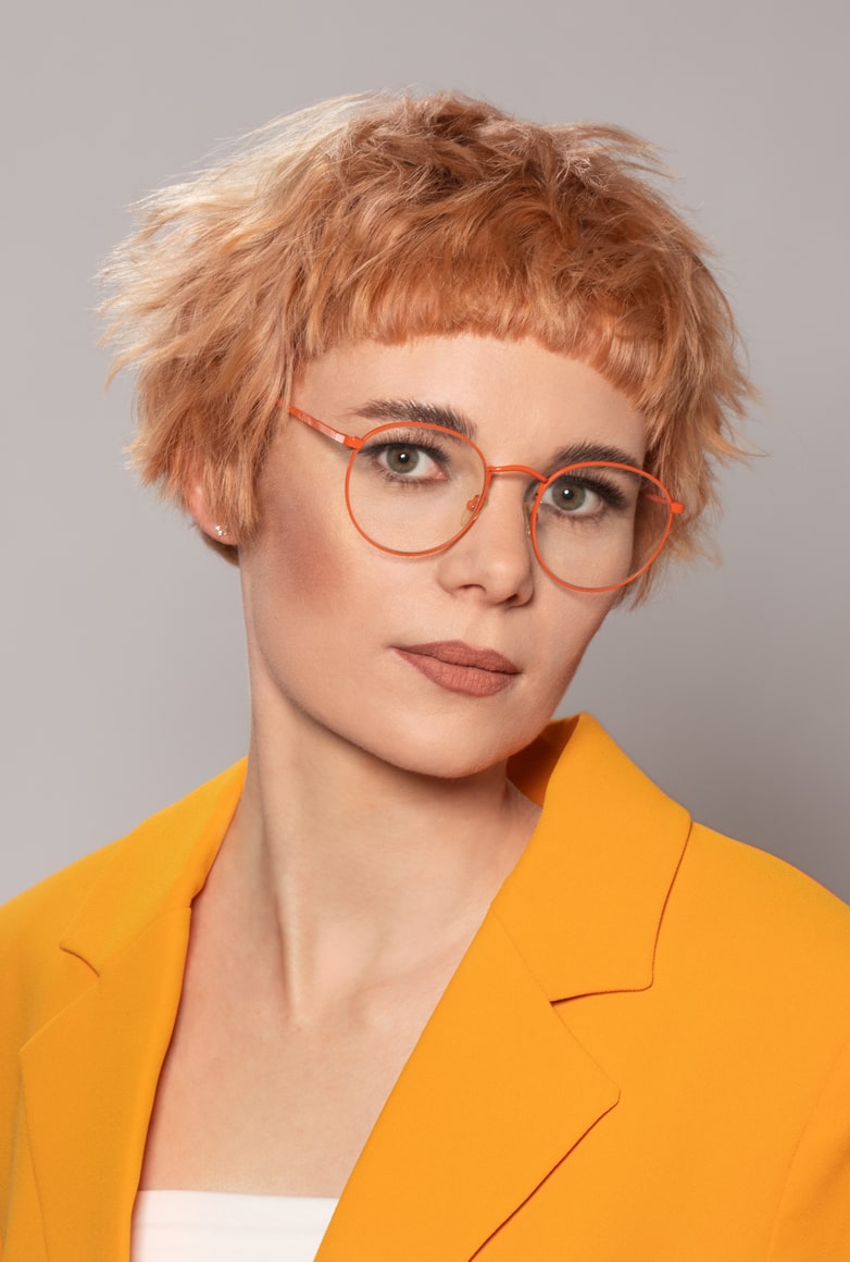 eine Frau mit einem extravaganten Pilzbob mit markantem Mikropony und Pastellkupfernen Haaren posiert in einem Orangenem Blazer für die Kollektion Sparkle von Unison